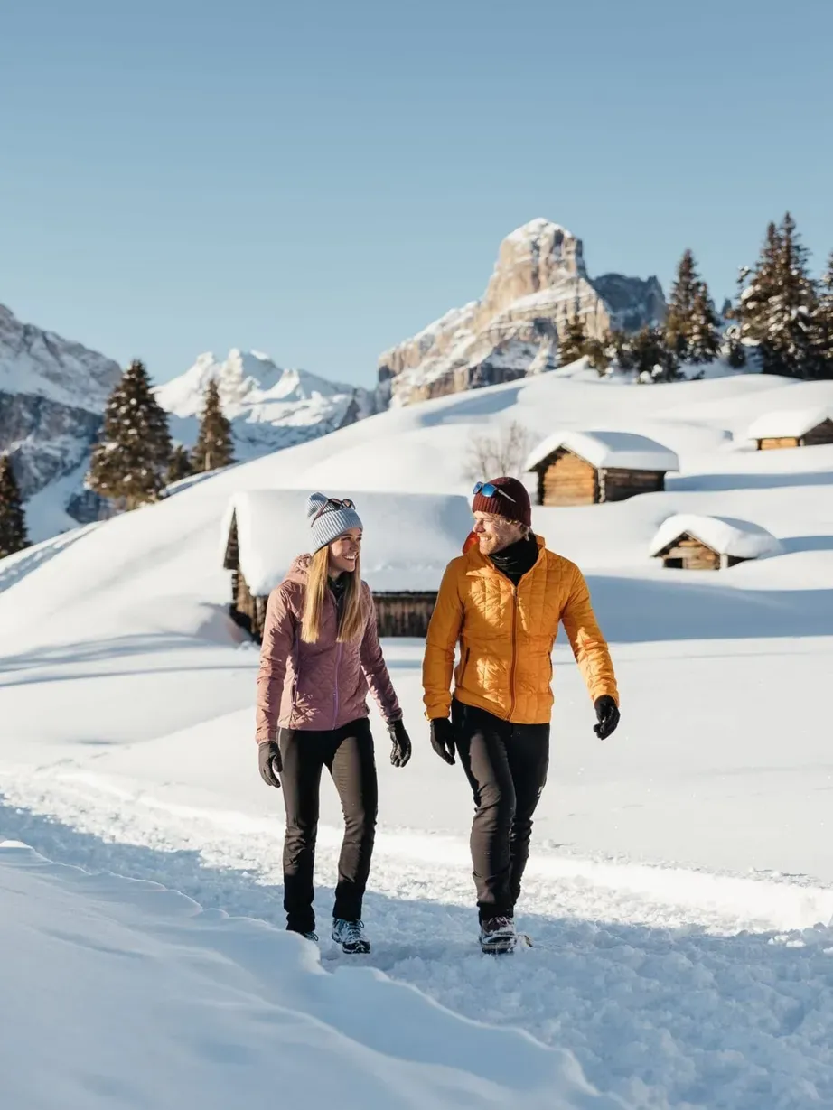 Two hikers walk along a snowy trail in Alta Badia, Dolomites, with pine trees, snow-covered slopes, and small wooden cabins under a clear blue sky.