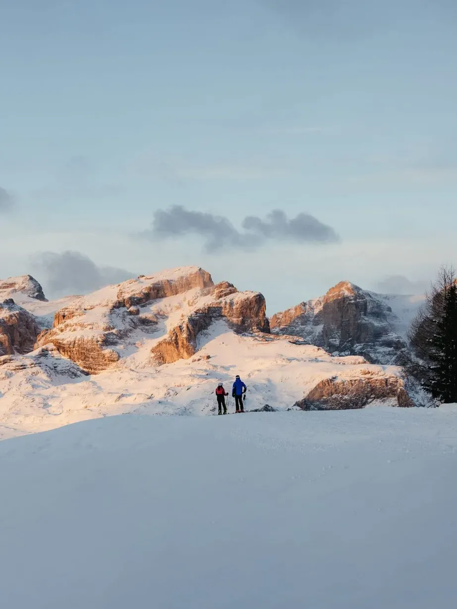 Two hikers on a snow-covered ridge in Alta Badia, Dolomites, with sunlit peaks behind and a few trees to the right under a pale blue sky.