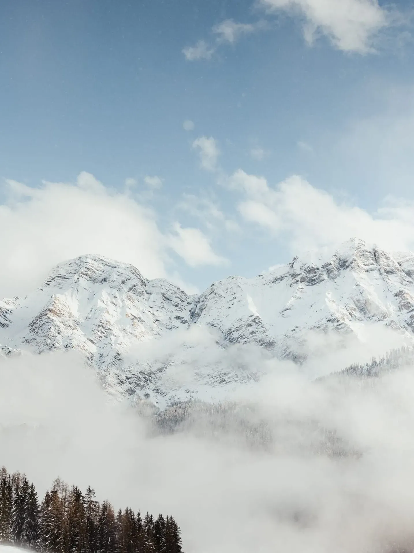 Snowy Dolomites peaks in Alta Badia rise above a pine forest, with wispy clouds drifting through the valley under a blue sky.
