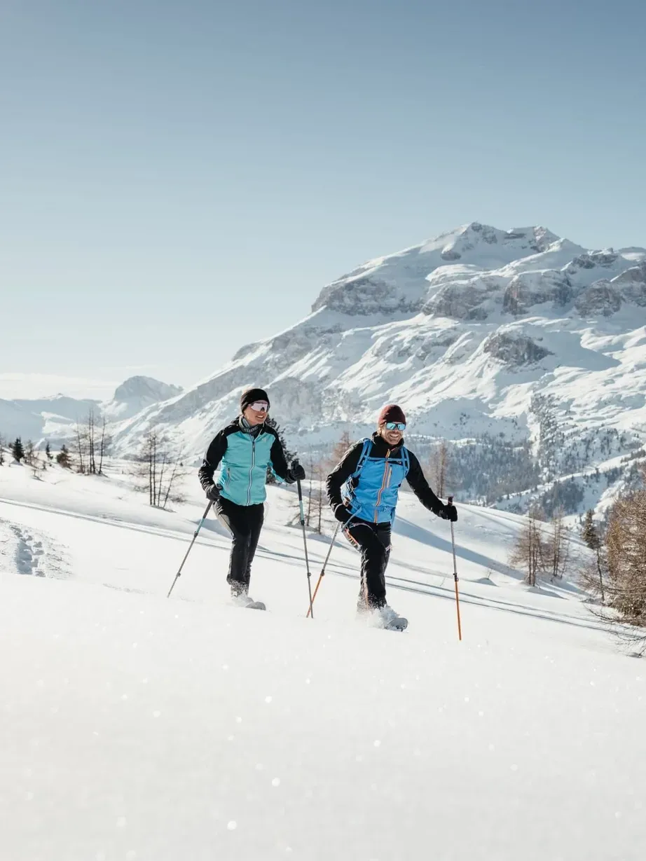 Two skiers in blue jackets ski uphill on a snowy slope, with snow-capped Dolomites and a clear blue sky behind them.
