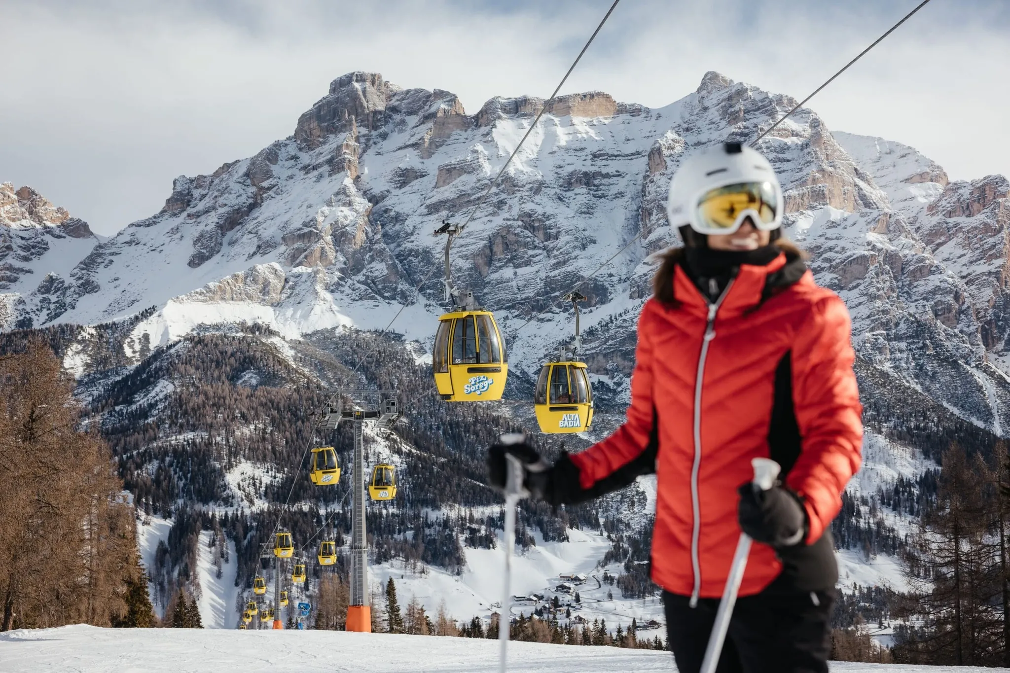 Snowy Dolomites with yellow gondola cabins along a mountain cable line; foreground shows a person in a red ski jacket, white helmet, and goggles holding ski poles.