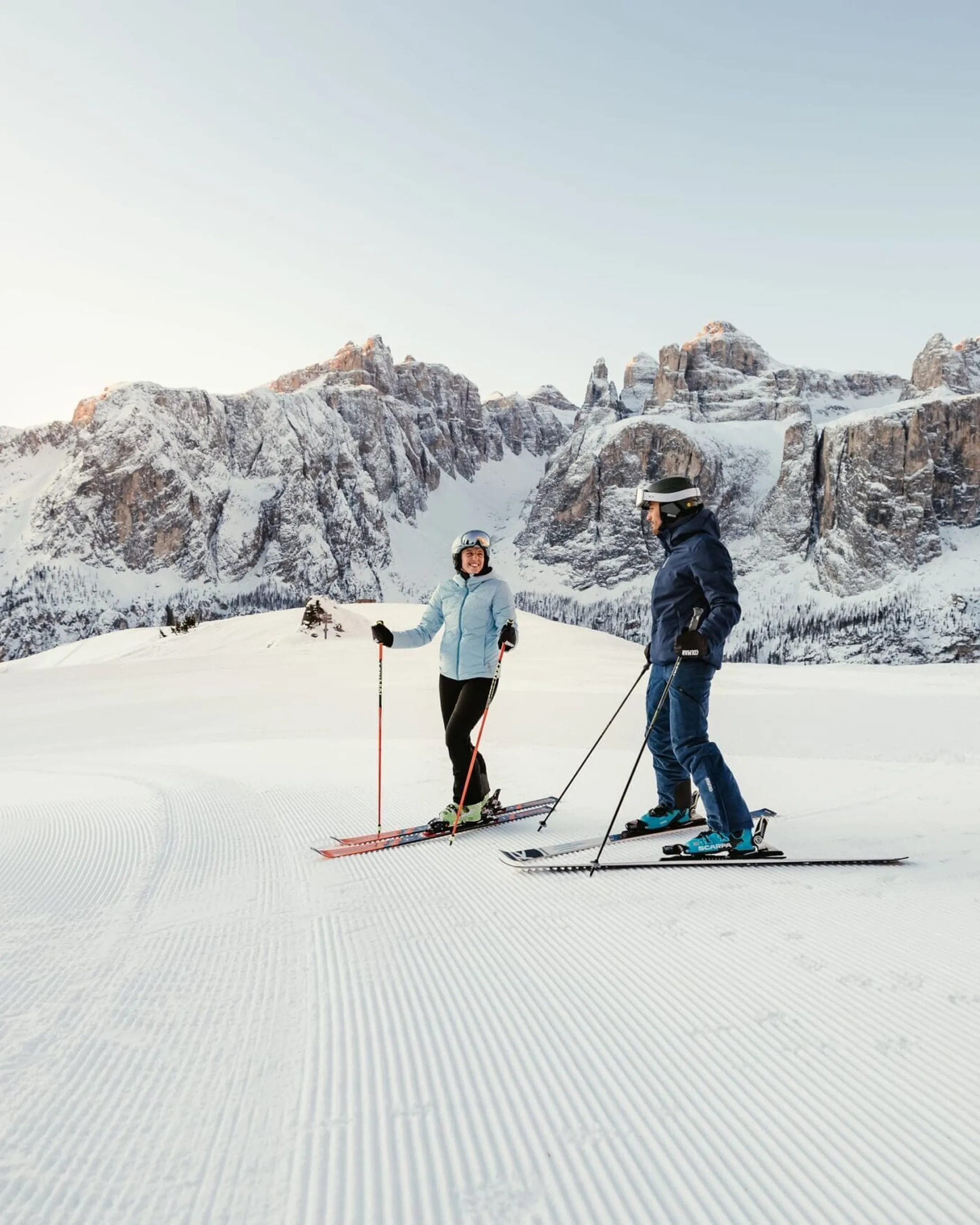 Zwei Skifahrende mit Helmen stehen auf einer präparierten Piste in Alta Badia, Dolomiten, bei sanftem Morgenlicht; dahinter majestätische, verschneite Felsformationen.