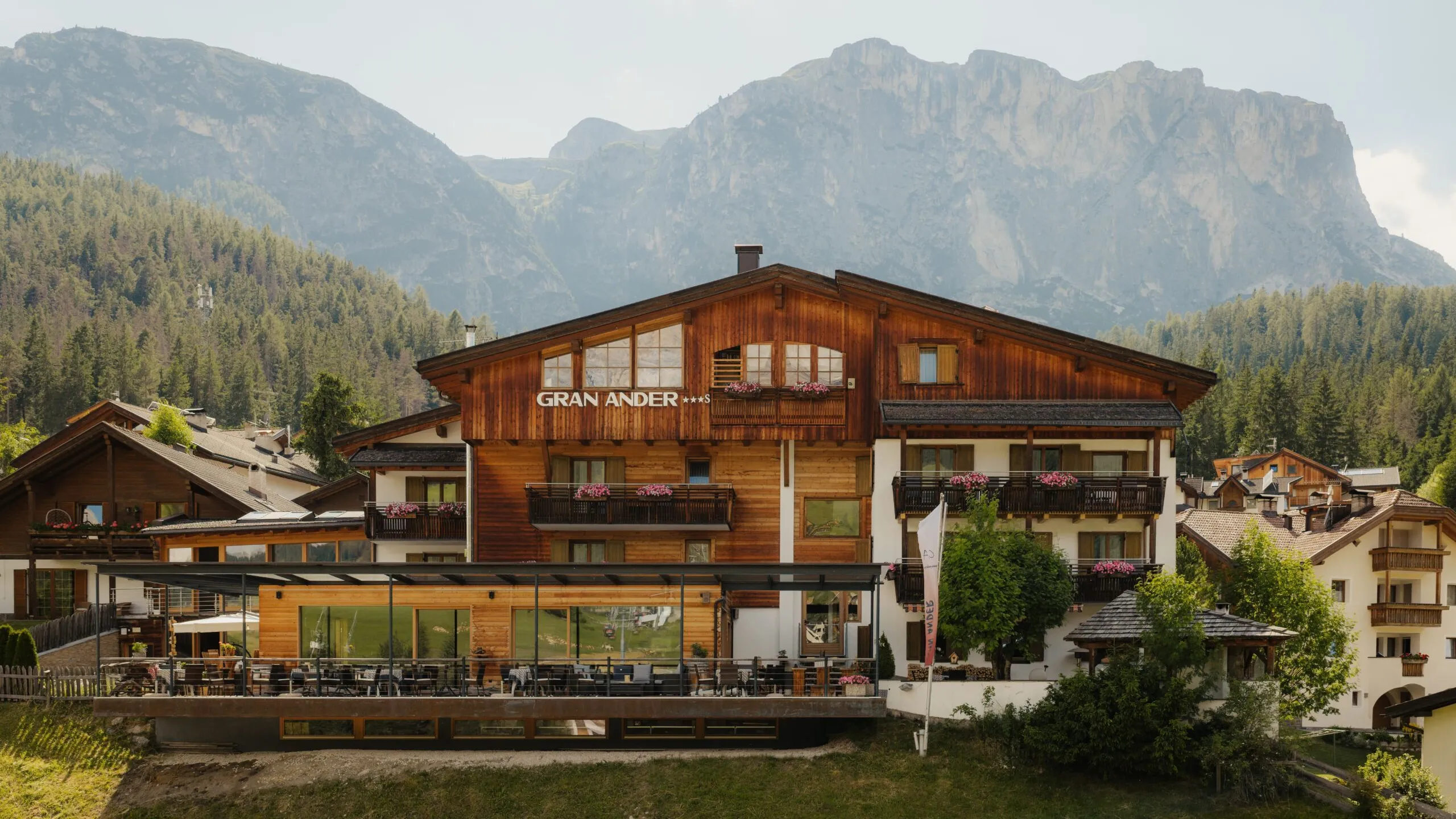 Hotel Gran Ander, chalet in legno con balconi fioriti e terrazza vetrata, ai piedi delle Dolomiti tra foresta alpina.