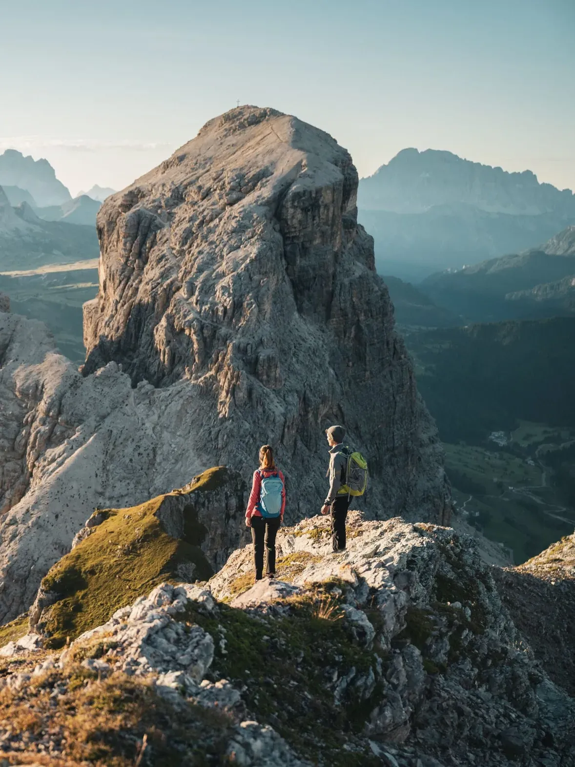 Two hikers with backpacks stand on a rocky ridge in the Dolomites, Alta Badia, surveying jagged peaks and a valley bathed in morning light.