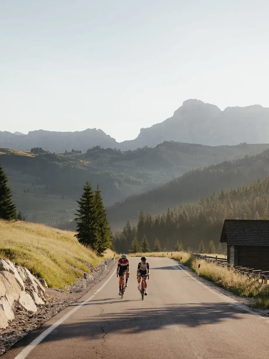 Two cyclists ride along a mountain road in the Dolomites, bordered by a rocky wall on the left and a wooden cabin on the right, with pine trees and distant peaks.
