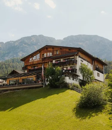 Hotel Gran Ander in Badia, Alta Badia, Dolomiten: mehrstöckiges Holzgebäude auf grünem Hang, Balkone mit Blumen, Bergkette und blauer Himmel.