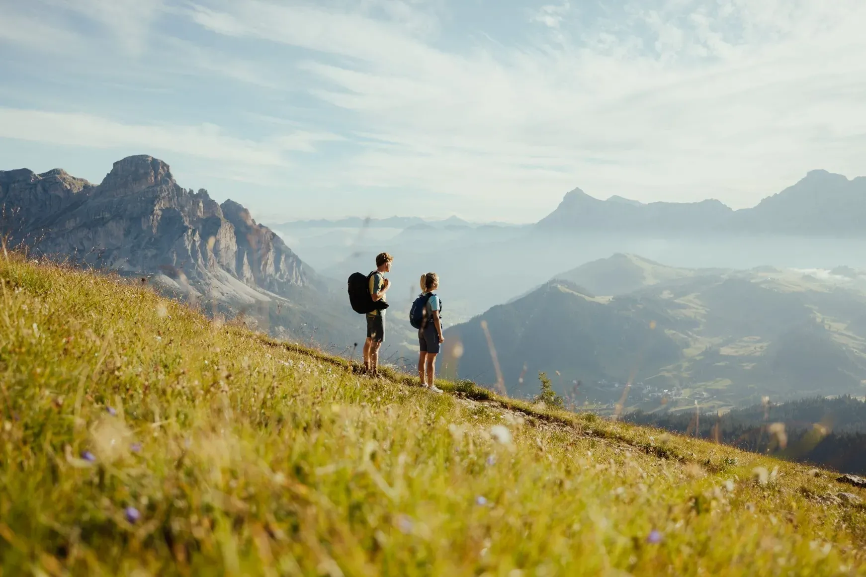 Two hikers with backpacks stand on a sunlit alpine meadow, gazing at rugged Dolomites peaks and a hazy valley under a blue sky.