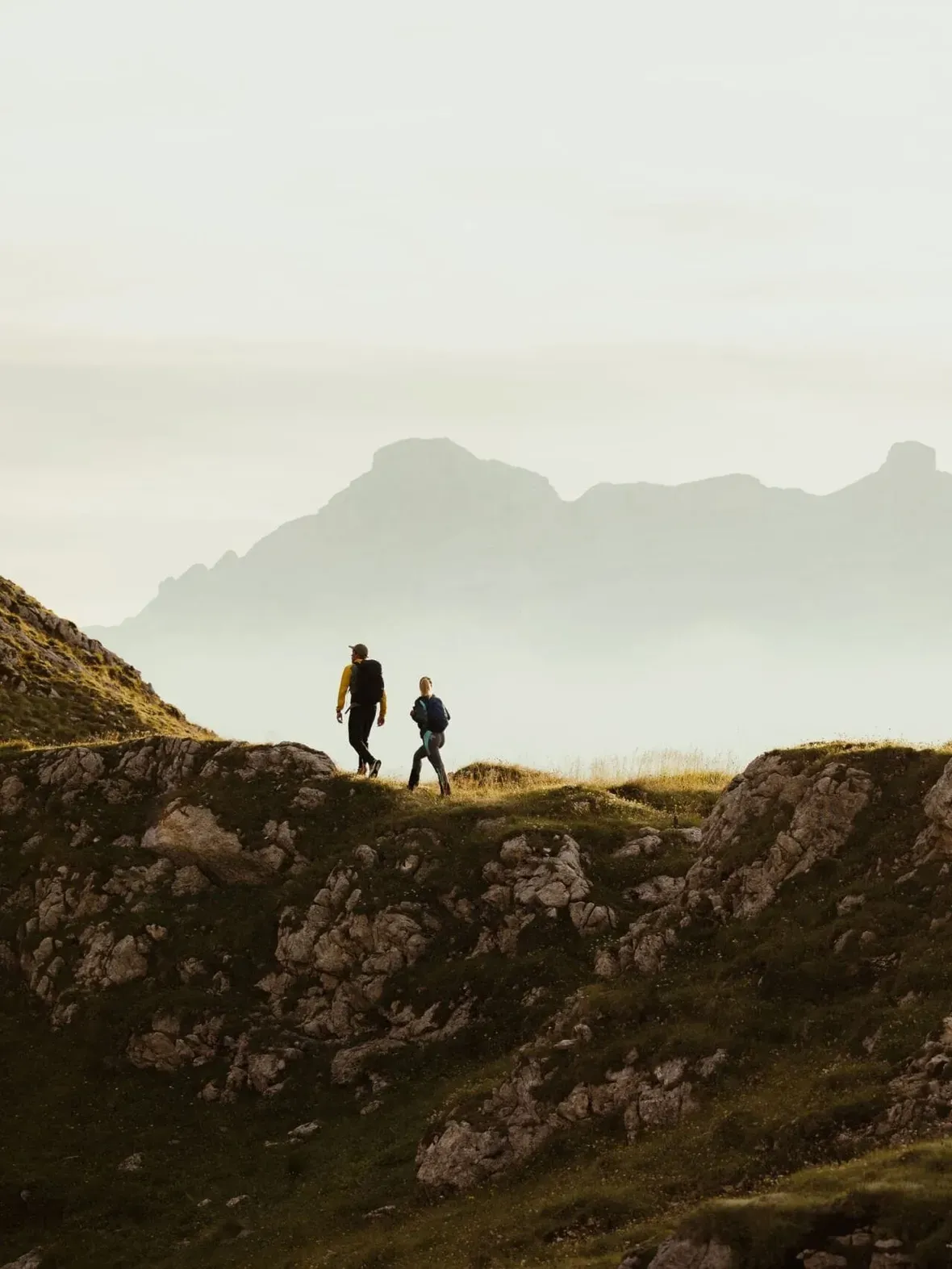 Two hikers walk along a rocky ridge in the Alta Badia Dolomites, with distant misty peaks and warm sunlight.
