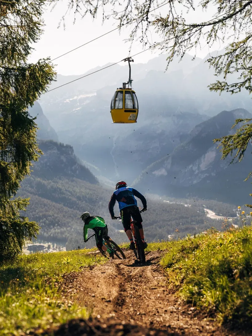 Two cyclists ride a dirt trail down a Dolomites mountainside; a yellow gondola hangs above as pine trees frame the scene and distant peaks loom.