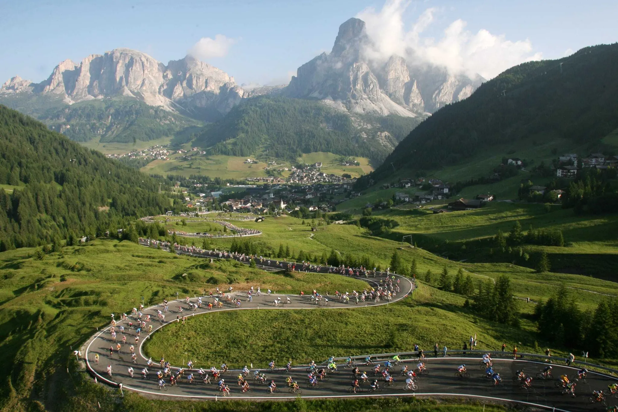 Winding road through green hills in Alta Badia, Dolomites, filled with cyclists; rugged peaks rise in the distance under a blue sky.