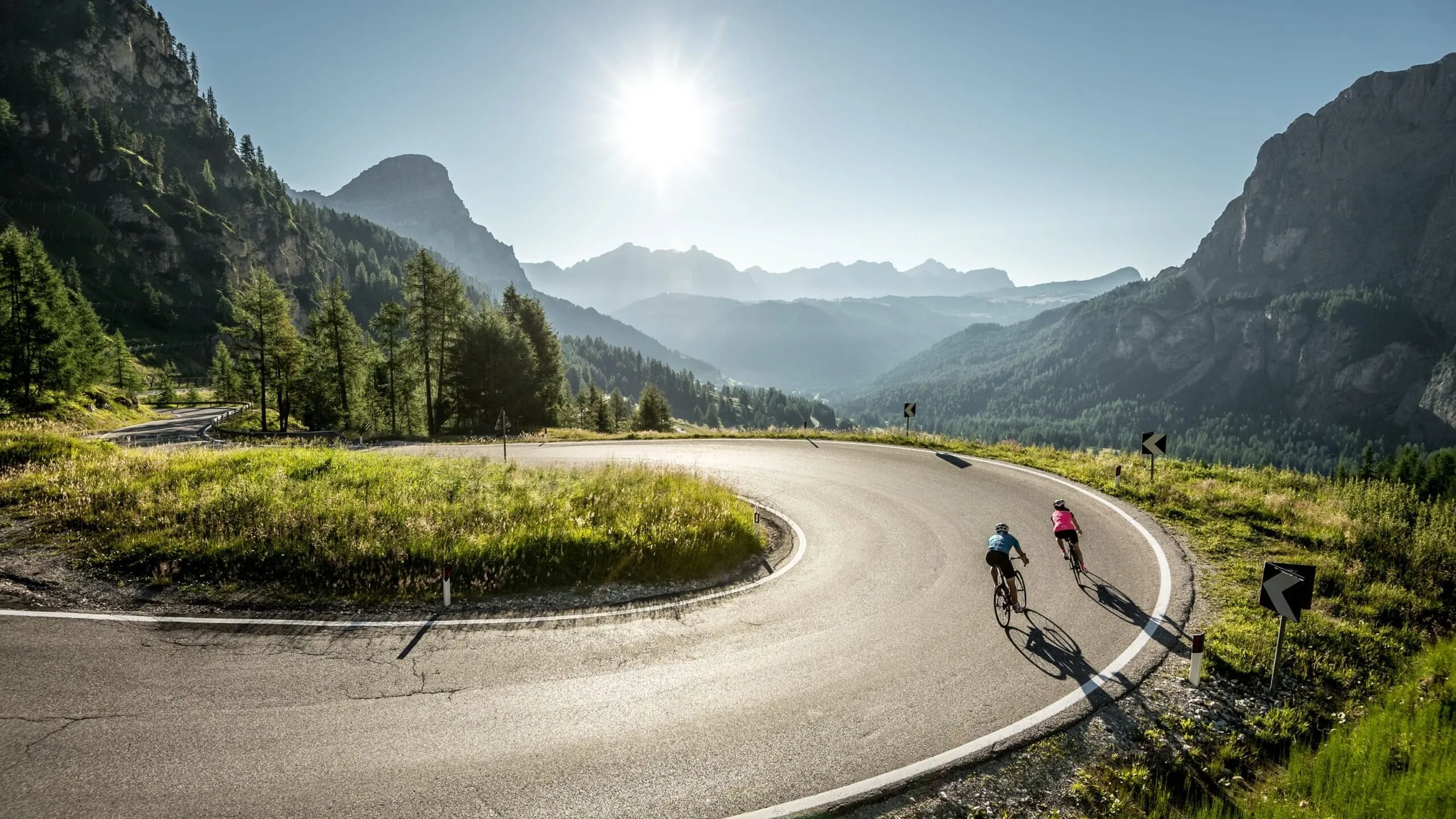 Zwei Radfahrende bewältigen eine Kurve auf einer bergigen Straße in Alta Badia, Dolomiten, bei strahlendem Sonnenschein vor alpine Felslandschaften.