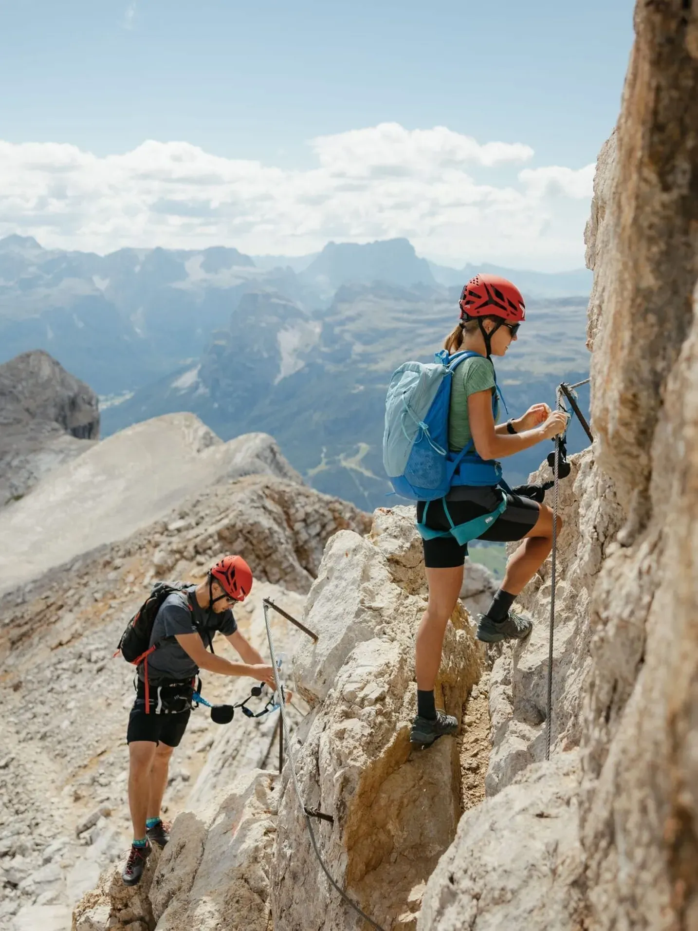 Two climbers with red helmets and harnesses ascend a narrow rocky ledge in Alta Badia, Dolomites, as valleys and peaks loom in the distance.