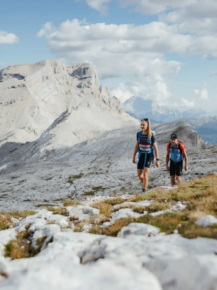 Two hikers on a rocky trail in the Dolomites, Alta Badia, with pale gray peaks, blue sky and scattered clouds.
