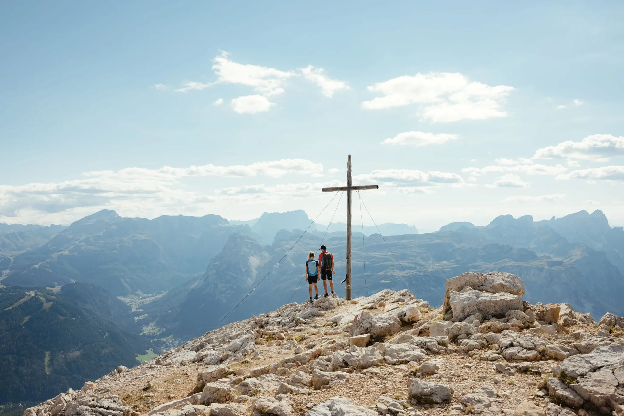 Two hikers stand on a rocky mountaintop beside a tall wooden cross, with the Dolomites sprawling below under a blue sky.