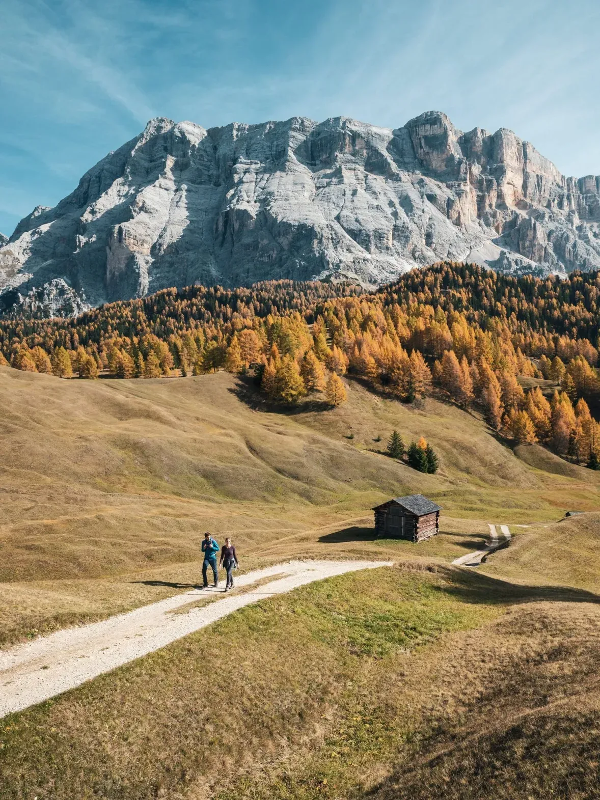 Autumn in Alta Badia, Dolomites: a dirt path winds across rolling meadows to a small wooden cabin, with golden trees and rugged peaks beneath a blue sky; two people walk.