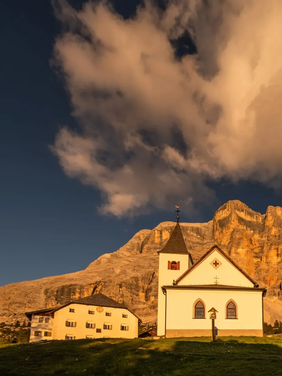 White church with a steeple on a grassy hill, Dolomites glowing gold behind under a blue sky with clouds in Alta Badia, Dolomites.