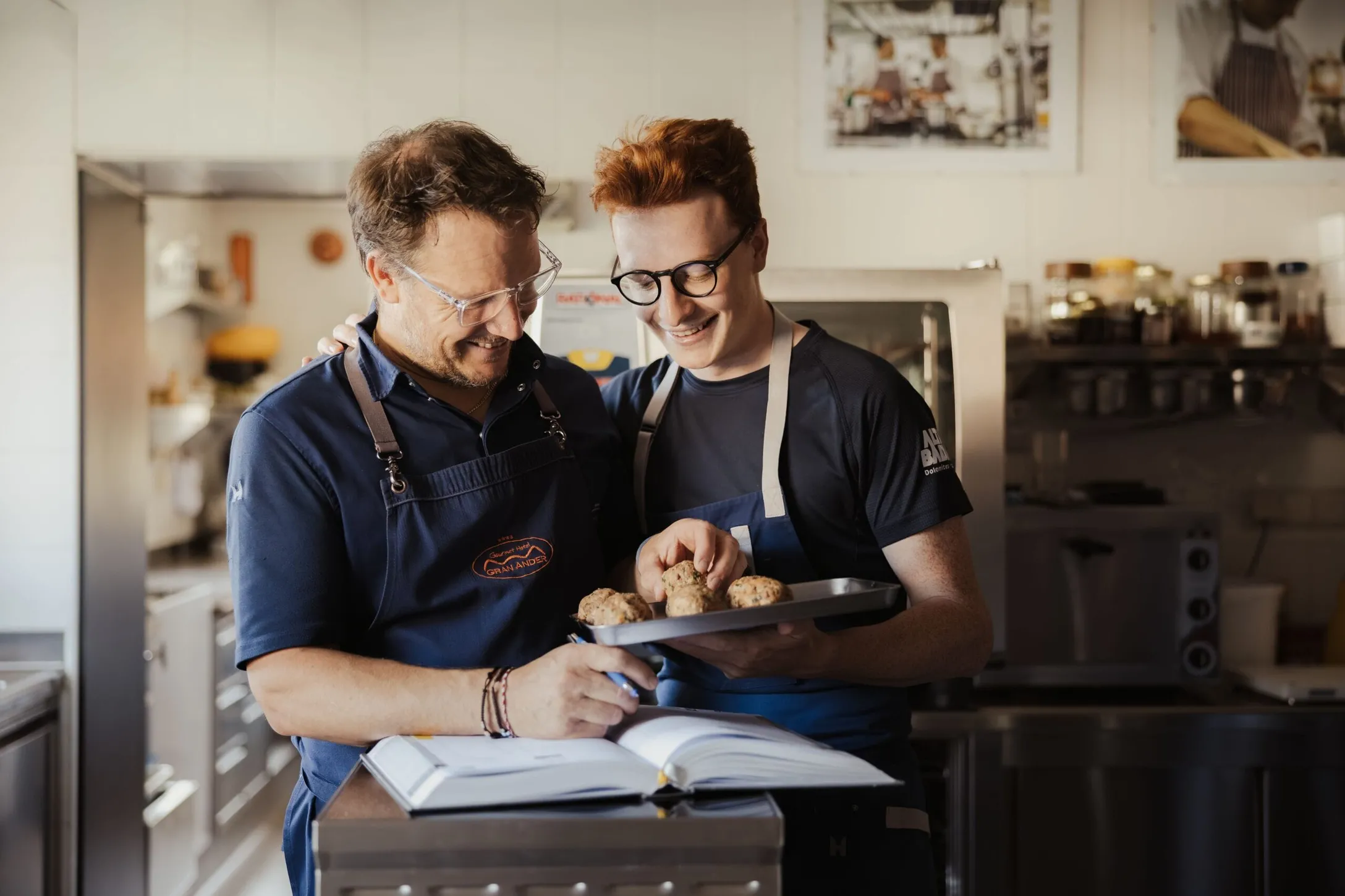 Maximilian und Andrea in einer Hotelküche (Gran Ander, Badia Dolomiten) mit Schürzen; sie halten ein Backblech mit Muffins, während ein offenes Kochbuch davor liegt.