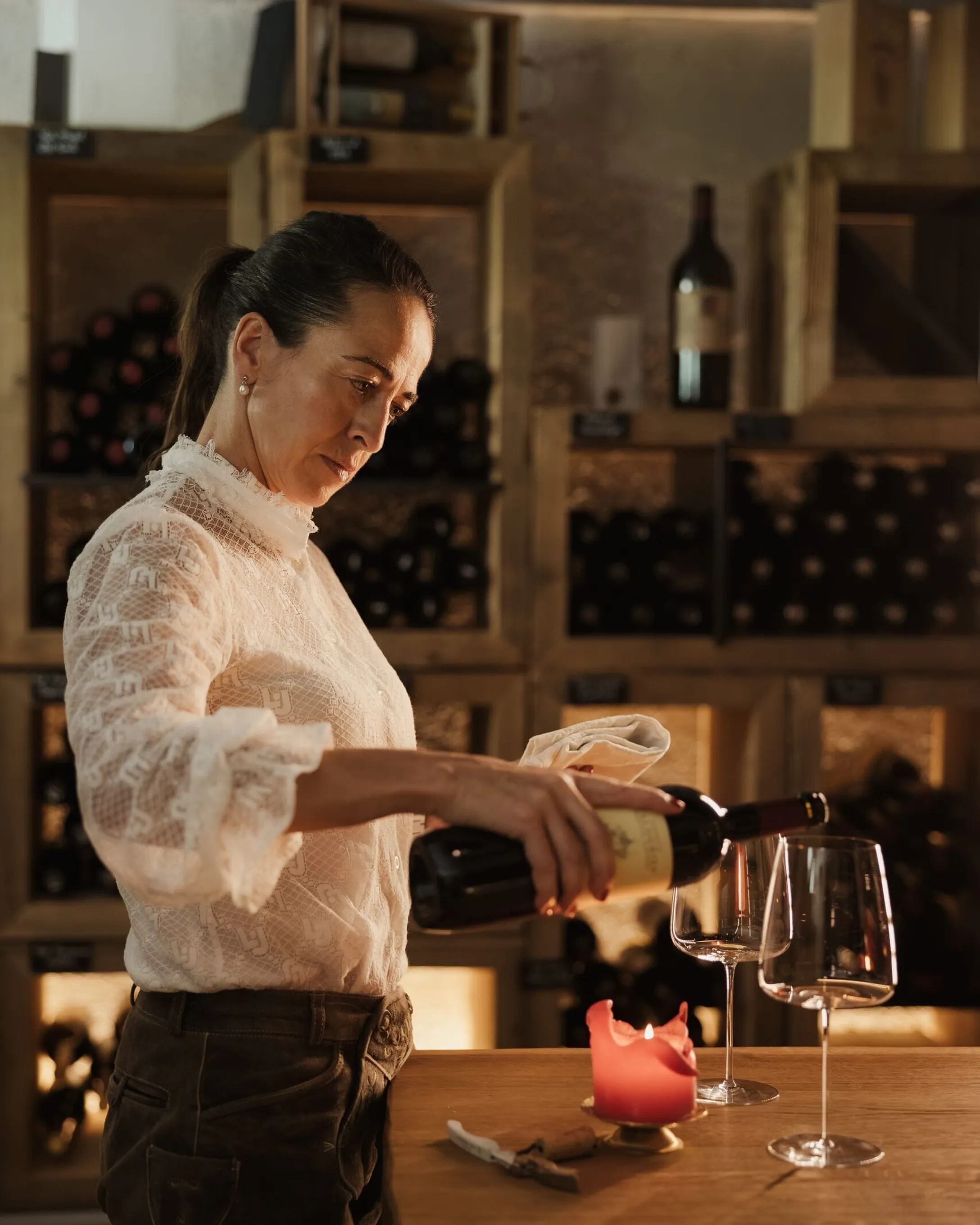 Evelin con camicia di pizzo bianca versa vino in un calice sul bancone di legno; sfondo scaffali pieni di bottiglie e una candela accesa.