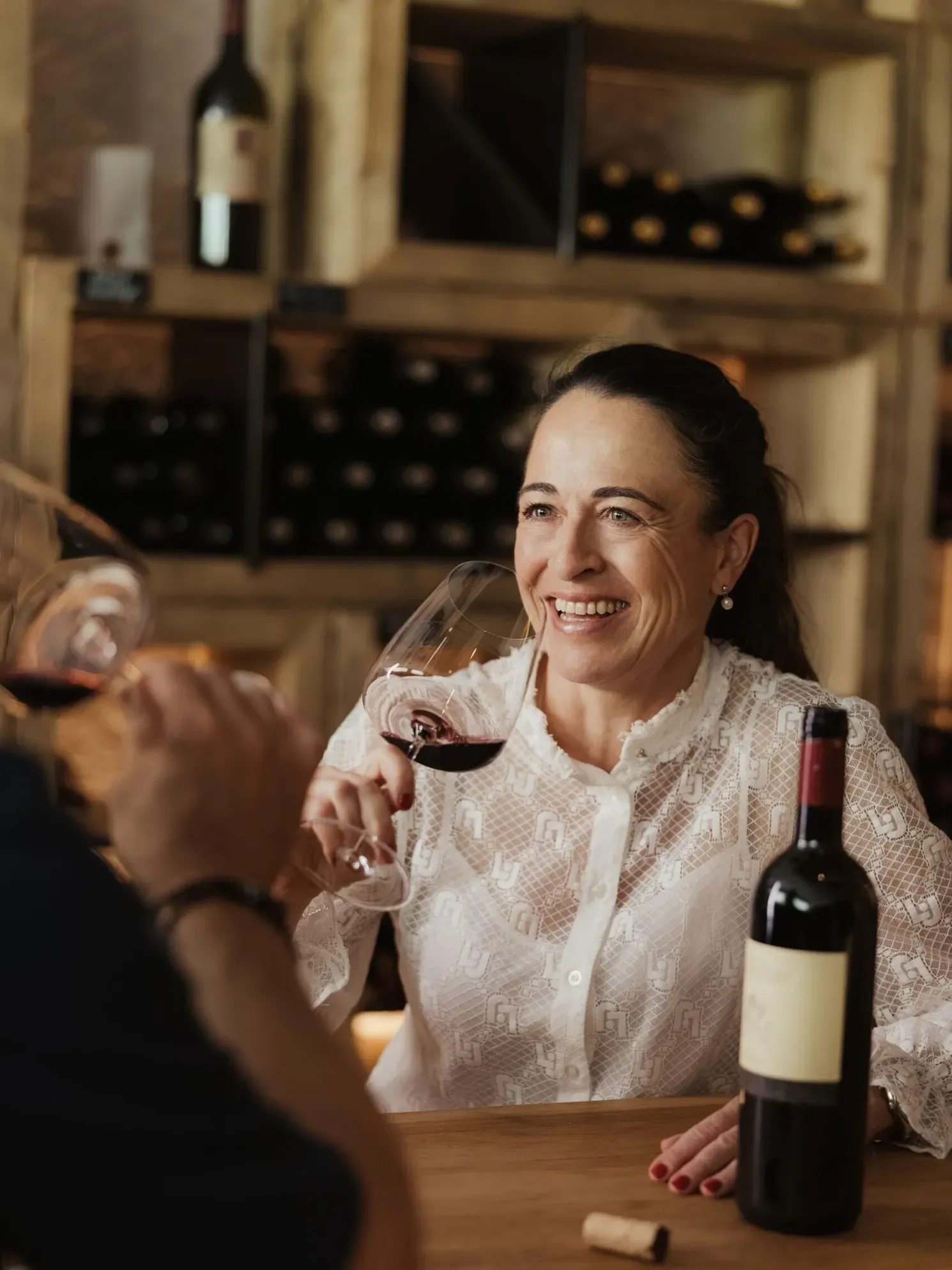 Evelin mit dunklem Haar in weißer Spitzenbluse lächelt, während Rotwein im Glas am Holztisch steht; Flasche, Korken und Weinregale im Hintergrund.