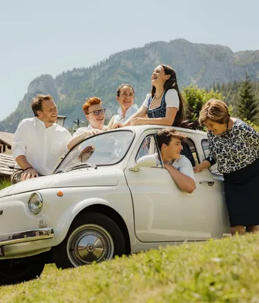 The Irsara family in a vintage white Fiat 500, happy on the meadow in Alta Badia, Dolomites, with the mountains in the background.