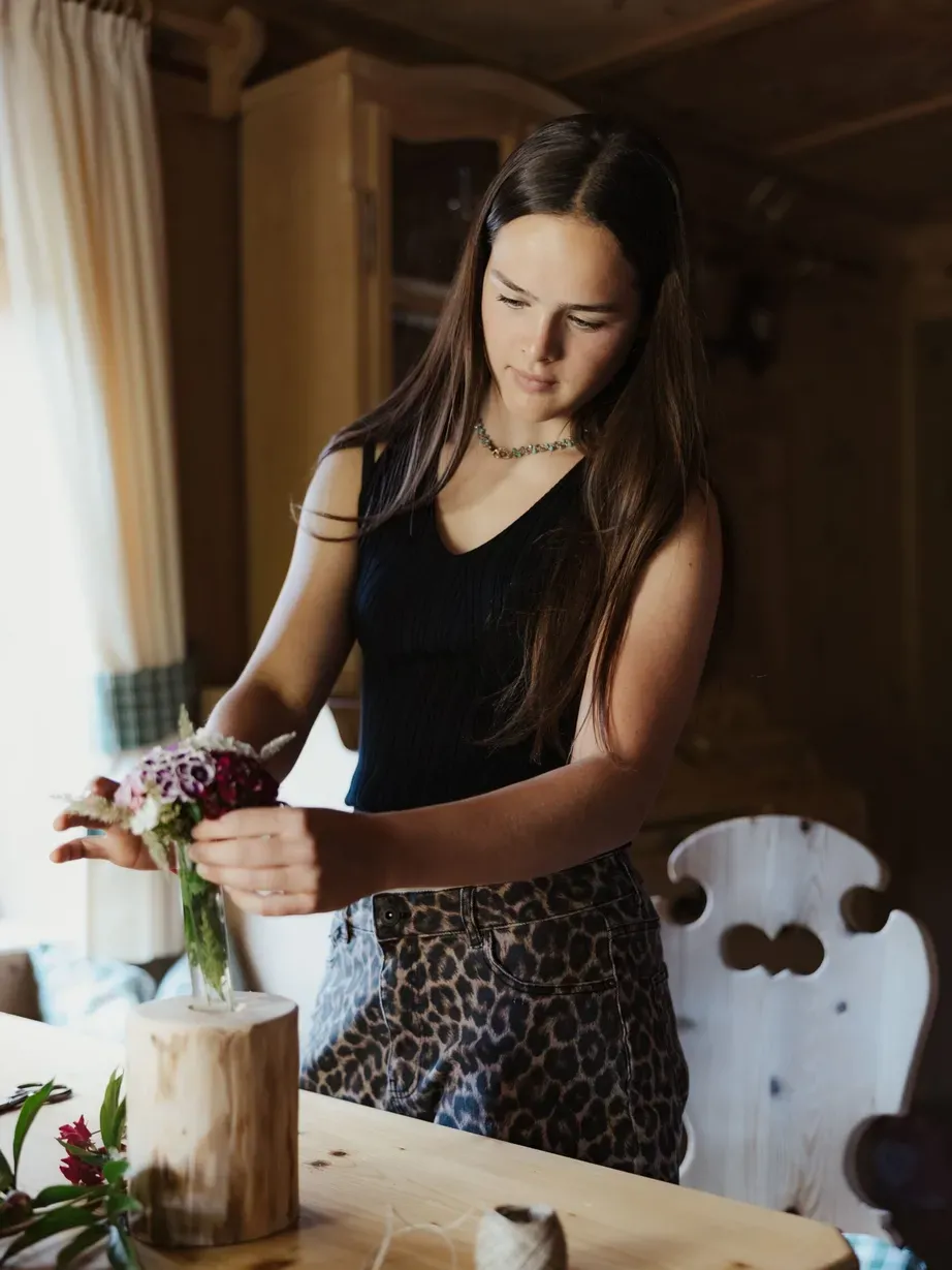 Lena con capelli lunghi scuri, top nero e gonna leopardata, sistema un bouquet in un vaso su un tavolo di legno.