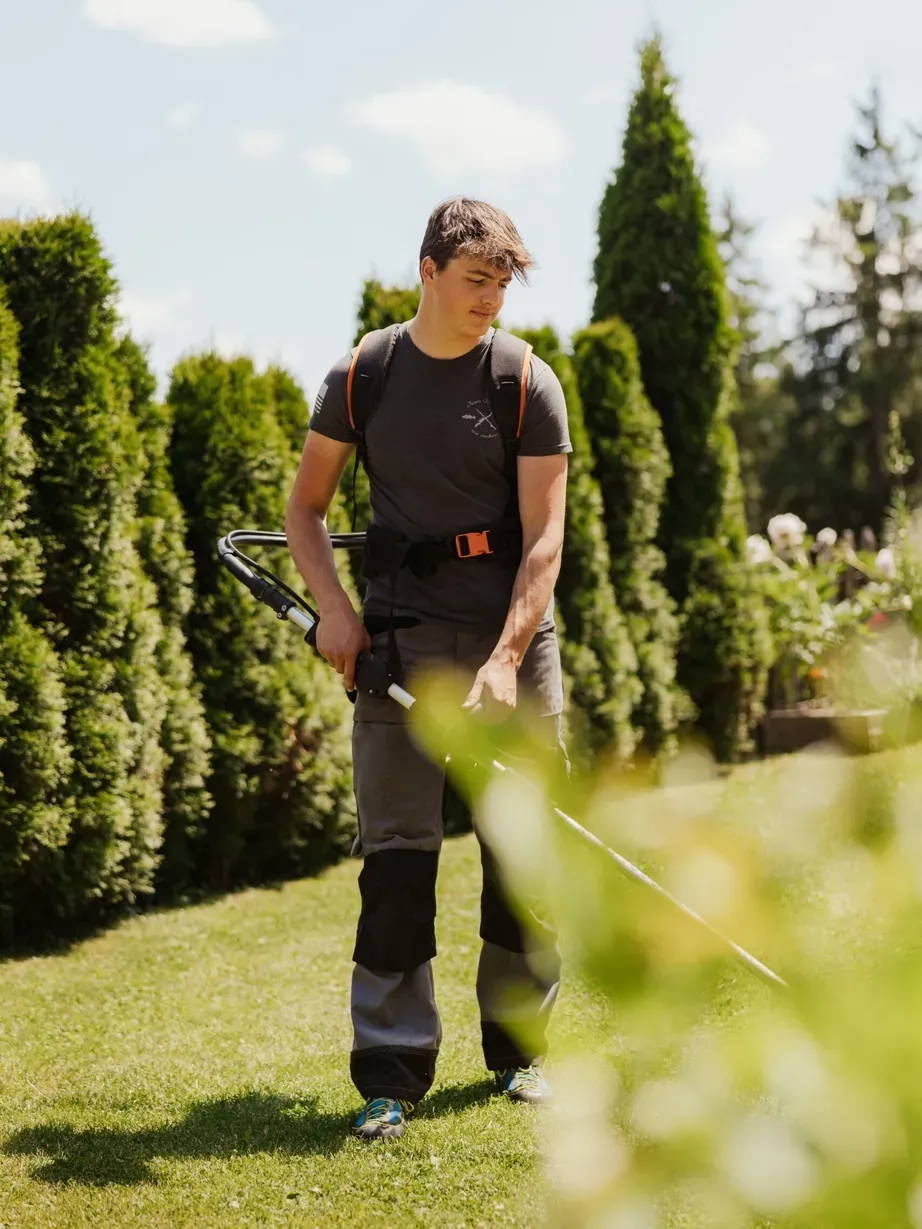 Tommy con zaino e abbigliamento da esterno usa un decespugliatore in un giardino soleggiato tra conifere in Alta Badia, Dolomiti.