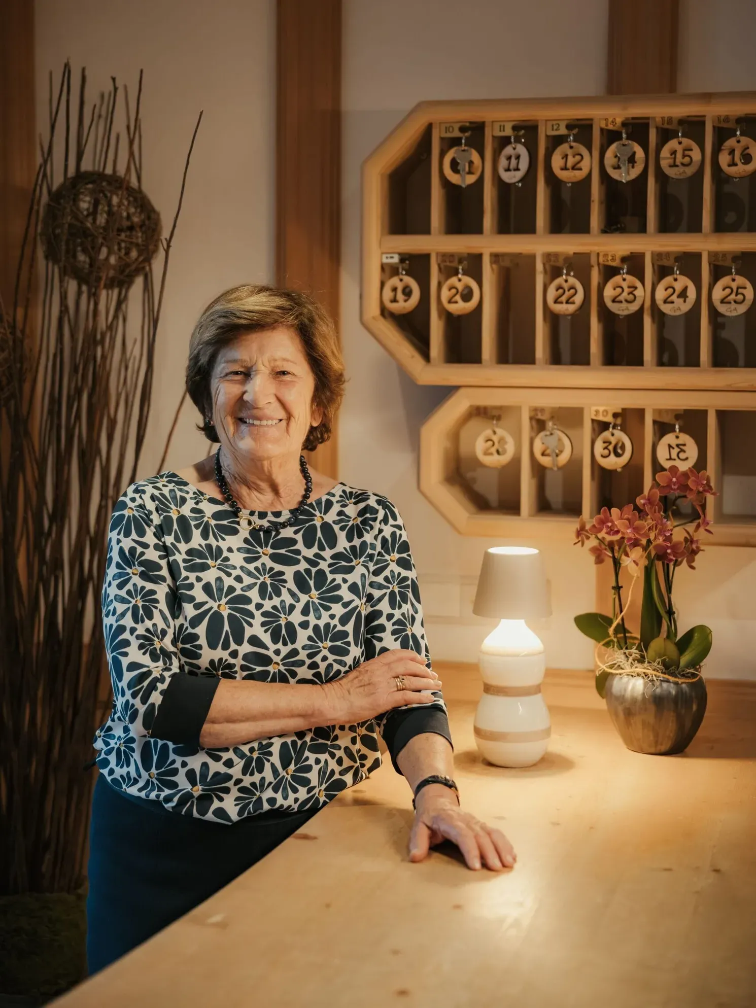 Rita sorridente dietro un bancone in legno nella hall dell'Hotel Gran Ander in Badia; sullo sfondo porta-chiavi in legno numerato, lampada accesa e vaso di orchidee.