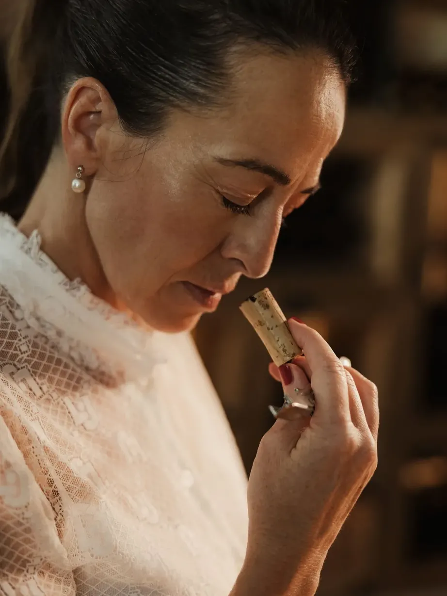 Evelin in primo piano, profilo sinistro, capelli raccolti, orecchini a perla, top bianco in pizzo, avvicina alle labbra un piccolo cilindro di cosmetico.