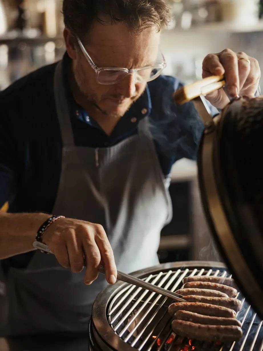 Andrea sta grigliando salsicce con una pinza su una griglia in cucina, tra vapore e luce calda.