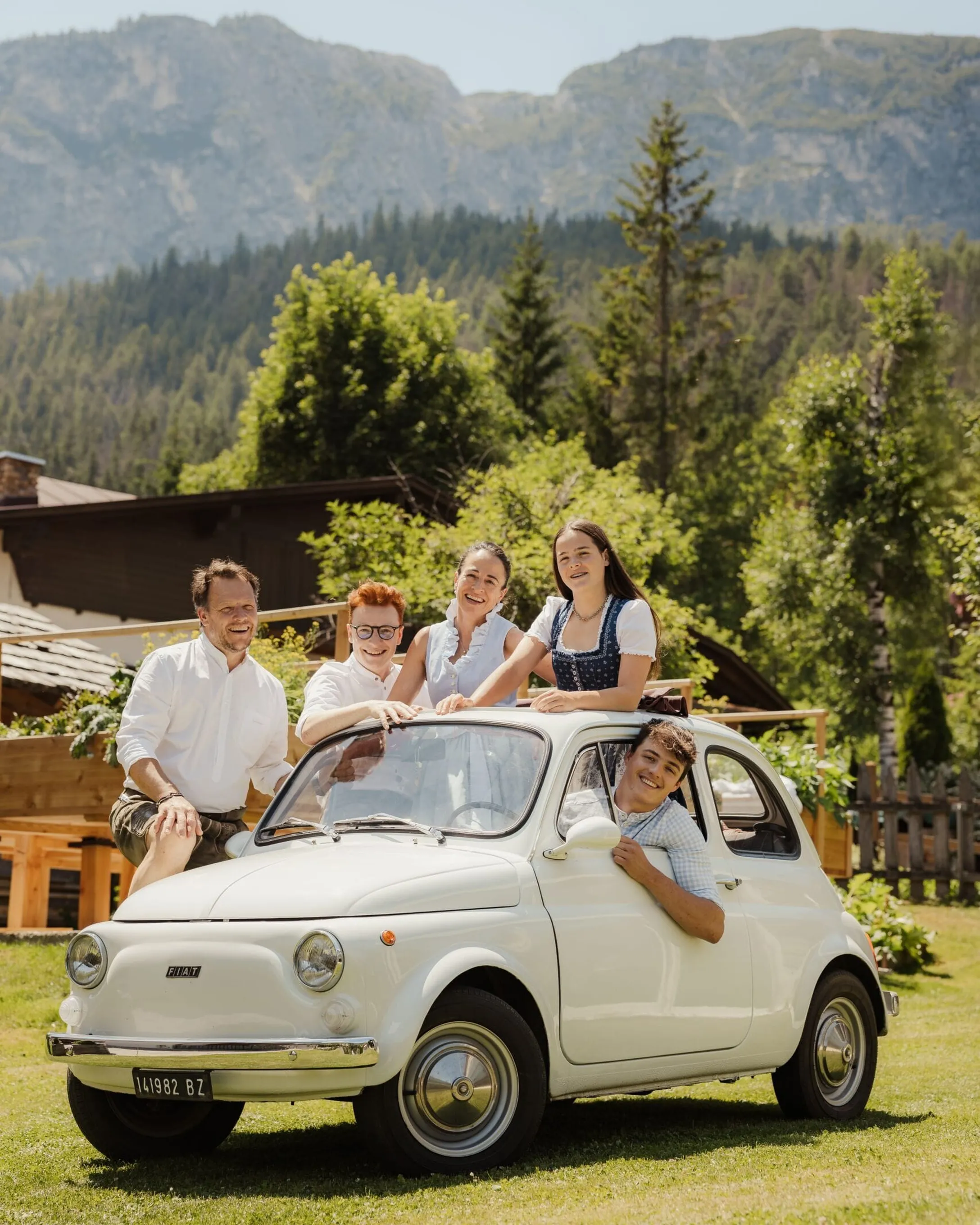Smiling members of the Irsara family lean against a vintage white Fiat 500 in a green meadow, with houses and the Dolomite landscape in the background.