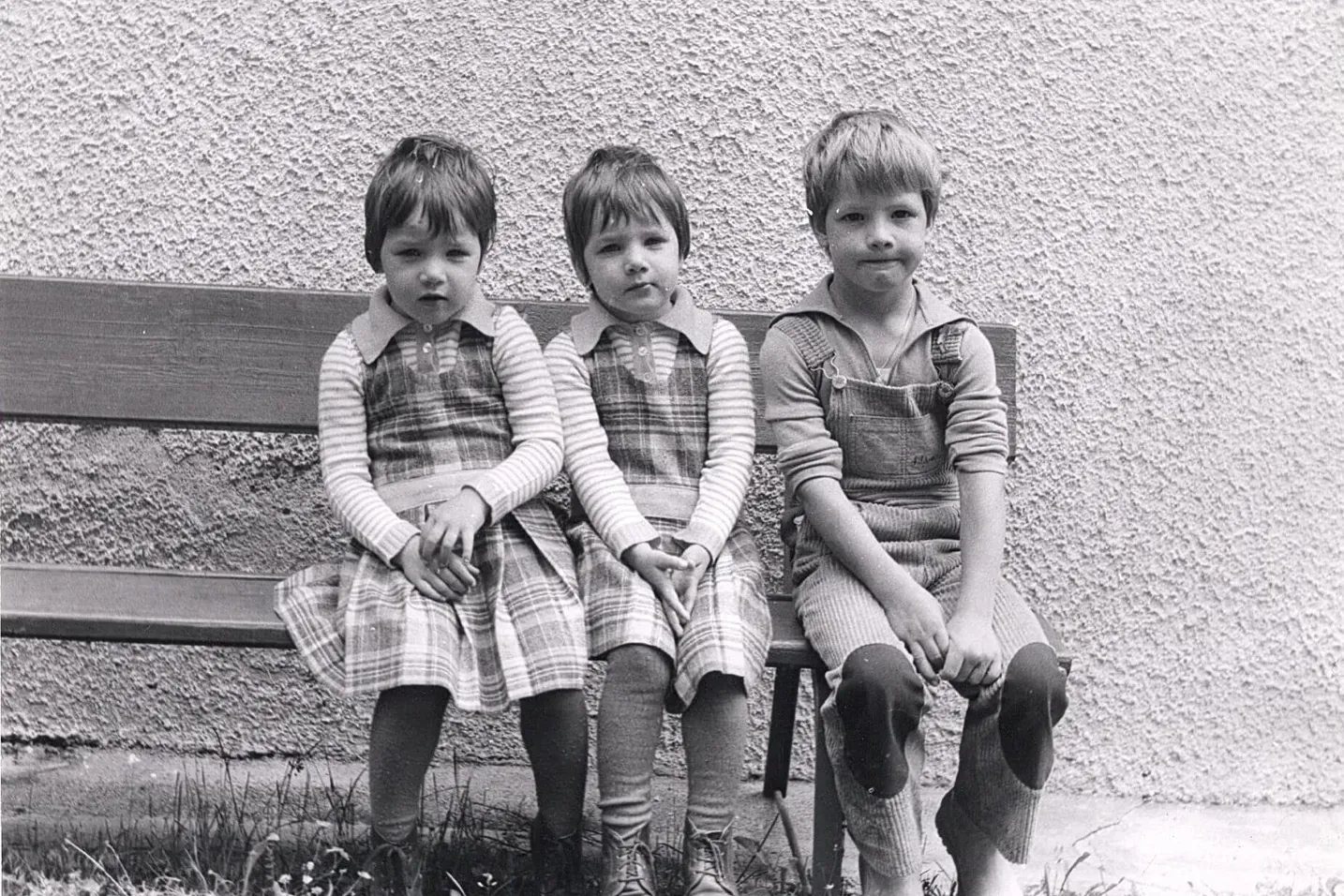 Tre bambini seduti su una panchina all'aperto davanti a una parete ruvida; due indossano abiti a quadri con maniche a righe, una in salopette.