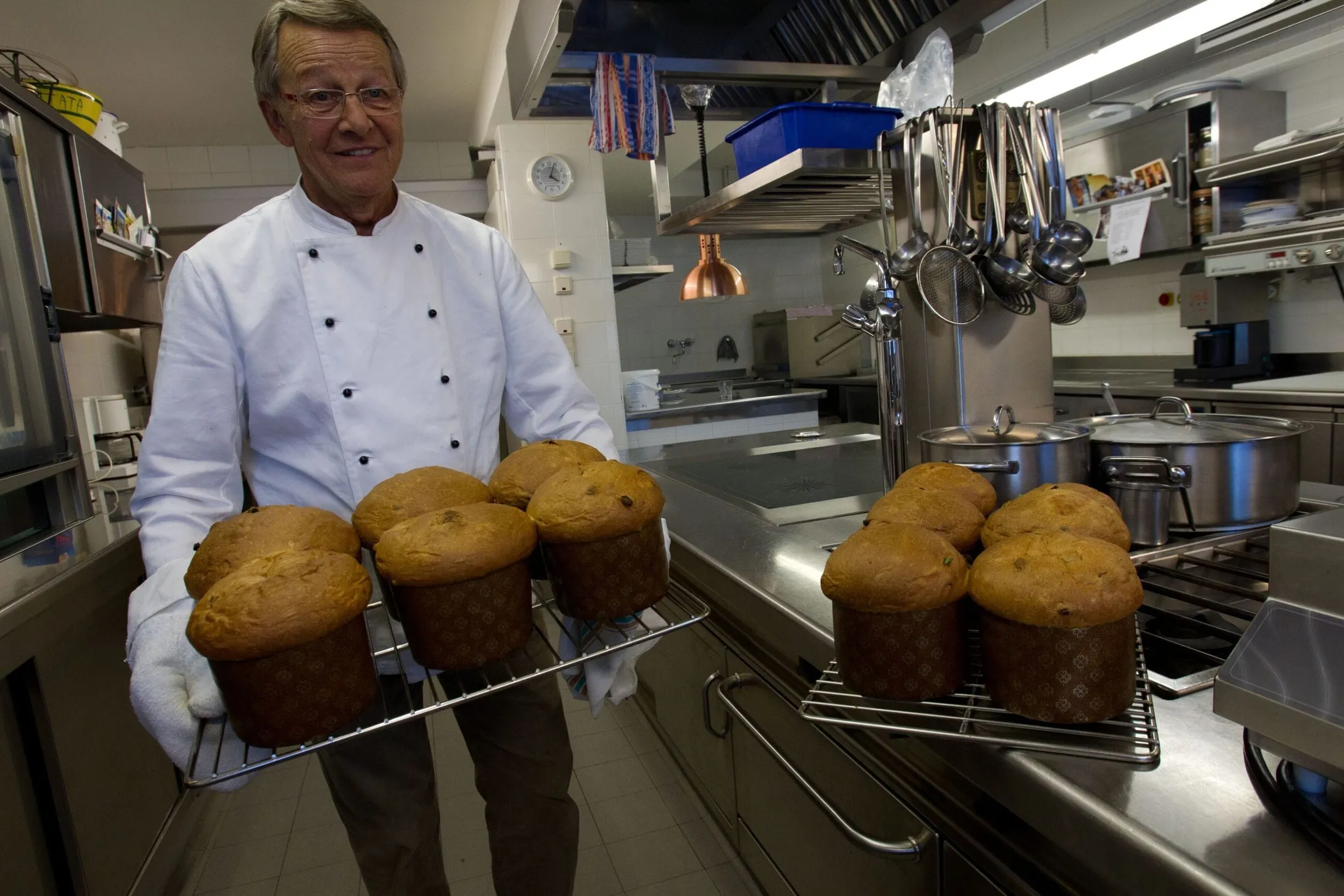 Chef in giacca bianca e guanti tiene due griglie di muffin dorati in una cucina professionale, con utensili appesi e pentole sullo sfondo.
