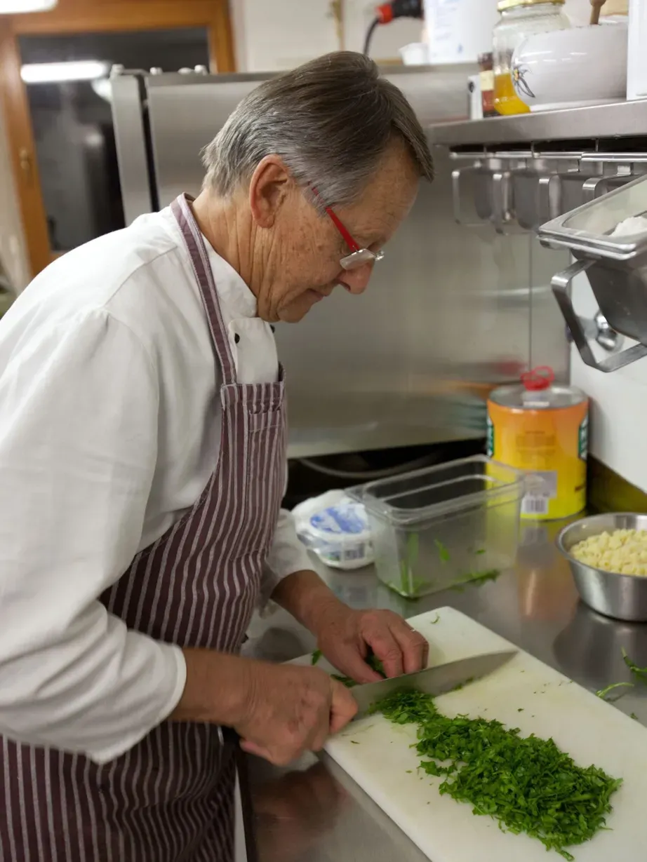 Una persona anziana in una cucina dell'Hotel Gran Ander a Badia: giacca bianca, grembiule a righe, taglia prezzemolo su tagliere bianco, attrezzature in acciaio sullo sfondo.