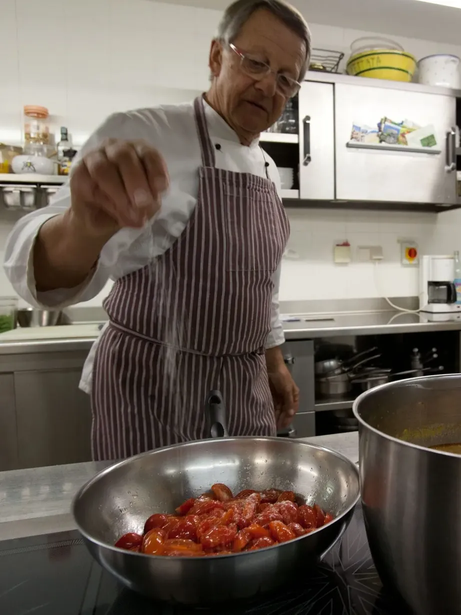 Una persona in cucina professionale, con grembiule a righe, porge una mano verso l'obiettivo, accanto a una grande pentola e a una ciotola di pomodori rossi.