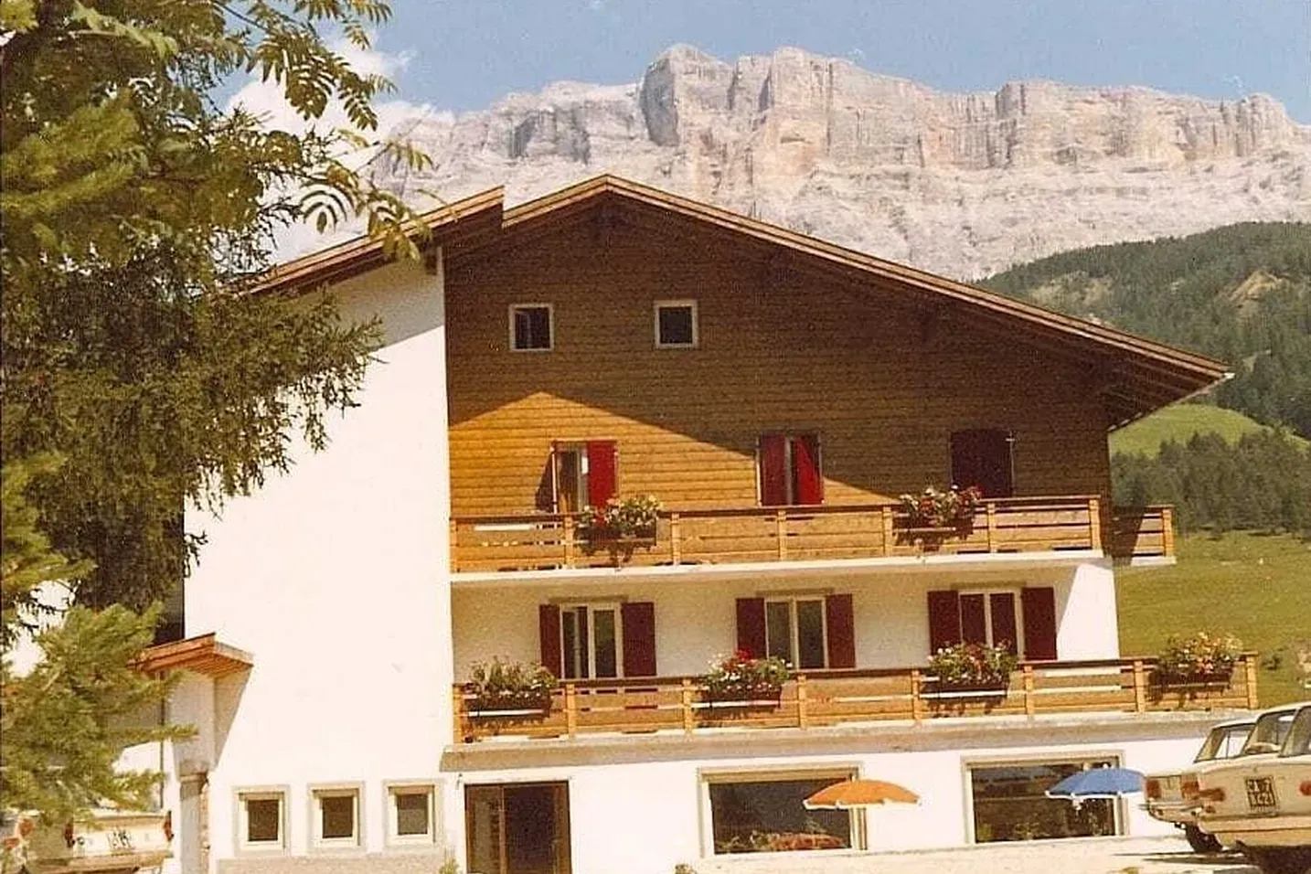 Hotel a tre piani, base bianca con piano superiore in legno, balconi fioriti, ombrelloni colorati, auto in parcheggio, Dolomiti sullo sfondo.