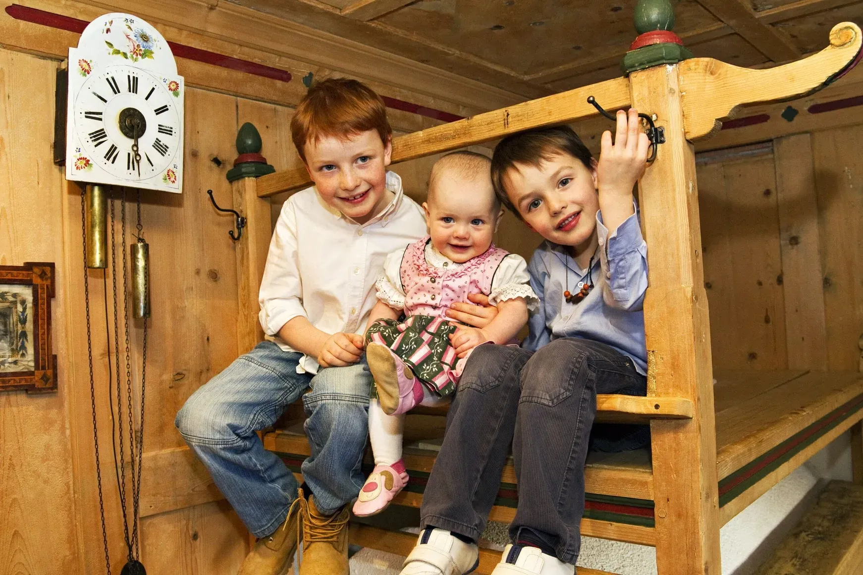Tre bambini seduti su una panca di legno in una stanza rustica; al centro un bebè, sfondo con orologio a pendolo e pareti di legno.