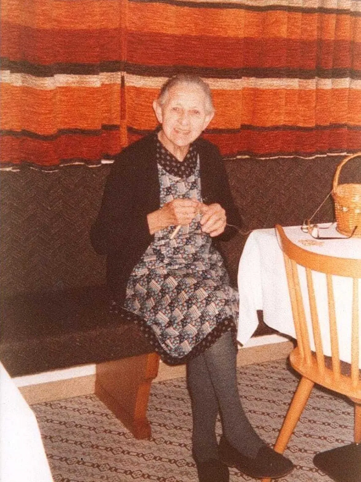 Persona anziana seduta su una panca, indossa vestito a fantasia, cardigan scuro e sciarpa; lavora a maglia accanto a tavolo apparecchiato, sfondo con tende arancioni.